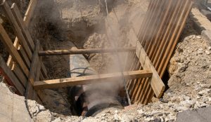 Trench under construction with a large pipe, wooden supports, and dusty atmosphere as workers install or inspect utilities.