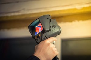 Handheld infrared camera in use, showing a colorful thermal image on the screen while held by a person near a wooden surface.